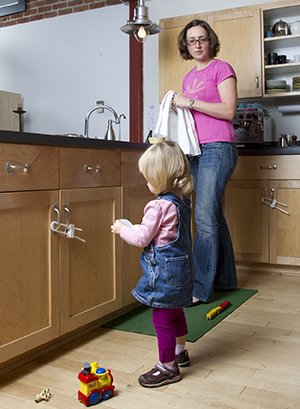 Woman watching toddler girl play in kitchen. Safety latches are on cabinets.
